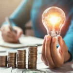 Лампочка в руке над монетами символизирует экономию на счетах за свет