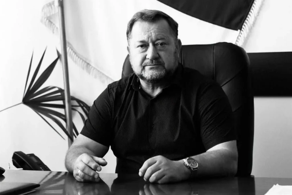 Чоловік у темній сорочці сидить за столом у кабінеті на тлі прапора