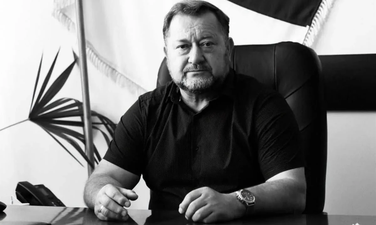 Чоловік у темній сорочці сидить за столом у кабінеті на тлі прапора