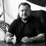Чоловік у темній сорочці сидить за столом у кабінеті на тлі прапора