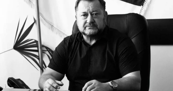 Чоловік у темній сорочці сидить за столом у кабінеті на тлі прапора