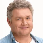 Чоловік із кучерявим сивим волоссям у блакитній джинсовій сорочці на білому тлі