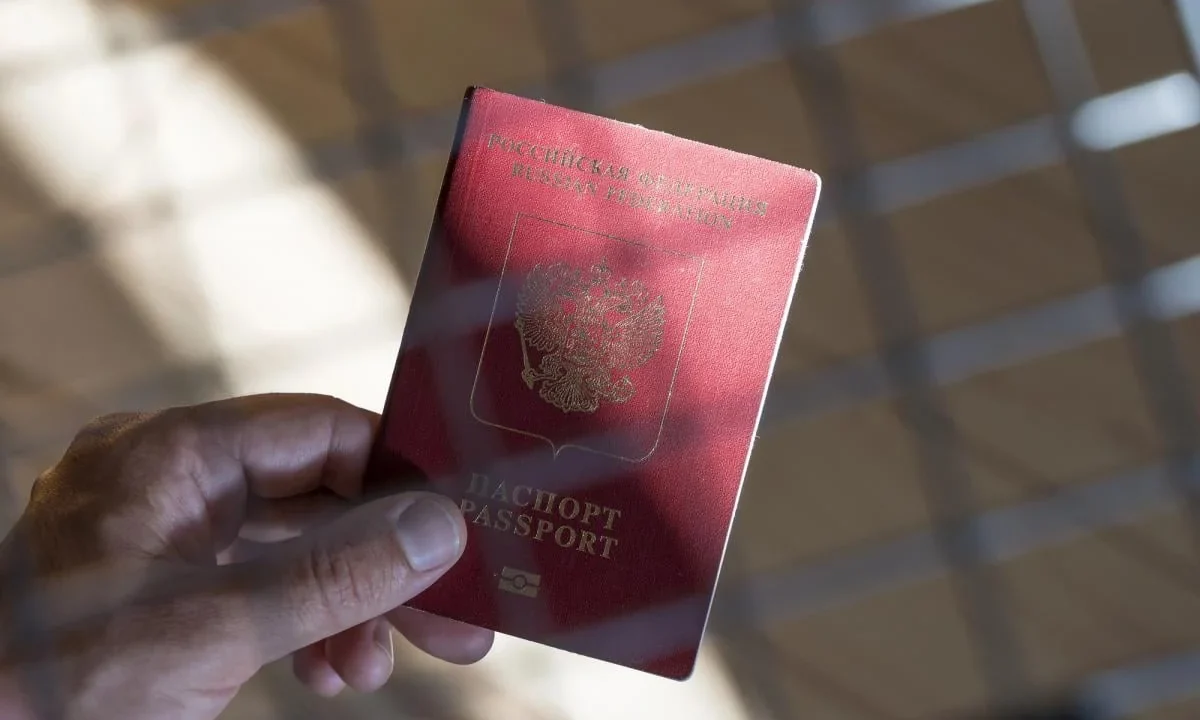 Рука тримає червоний паспорт Російської Федерації на тлі віконної сітки