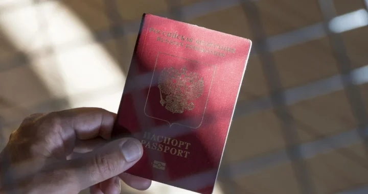 Рука тримає червоний паспорт Російської Федерації на тлі віконної сітки