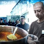 Чоловік розливає суп із великого казана, позаду група людей у дворі в’язниці