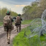 Два військових у камуфляжі йдуть ґрунтовою дорогою повз колючий дріт