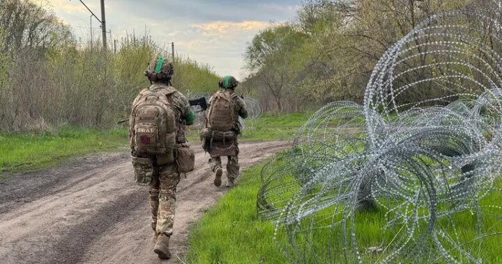 Два військових у камуфляжі йдуть ґрунтовою дорогою повз колючий дріт