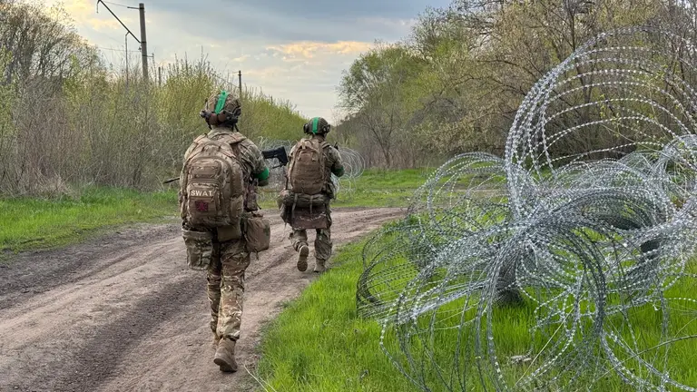 Два військових у камуфляжі йдуть ґрунтовою дорогою повз колючий дріт